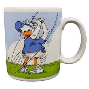 May include: White ceramic mug featuring a cartoon duck in a blue golf outfit swinging a golf club. The mug has a white handle and a light blue background with green grass. The duck is wearing a blue hat and shirt.