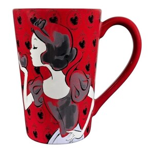 May include: A red ceramic mug with a Snow White design. The mug has a red handle and a repeating apple pattern on the background. The Snow White illustration is in black, white, and red, holding a dark apple.