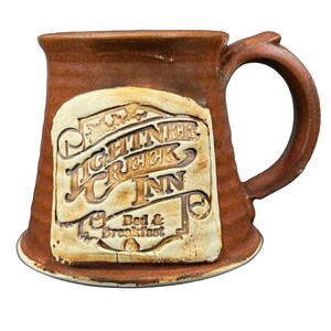 Peut inclure: Une chope en céramique marron avec une grande anse. La chope présente un emblème rectangulaire de couleur crème avec les mots "Lightner Creek Inn Bed & Breakfast" dans une police rustique. La chope a une surface texturée.