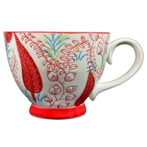 May include: White ceramic teacup with a red base and rim, featuring a floral design in red, blue, and green. The cup has a large handle and a decorative embossed pattern. A stylish addition to any kitchen.
