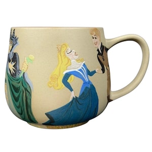 Könnte beinhalten: Cremefarbene Keramiktasse mit Figuren aus Dornröschen. Die Tasse zeigt Prinzessin Aurora in einem blauen Kleid, Prinz Phillip und Maleficent. Die Tasse hat einen großen Henkel und eine weite Öffnung.