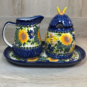 May include: A ceramic creamer and sugar bowl set with a matching tray. The set features a blue and white floral design with sunflowers and bees. The creamer has a handle and spout, and the sugar bowl has a lid with a bee-shaped handle.