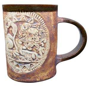 Puede incluir: Una taza de cerámica cilíndrica con un esmalte marrón moteado y un borde oscuro. El exterior presenta un relieve prominente de un león dentro de un borde circular, rodeado de patrones geométricos triangulares en un tono beige claro.