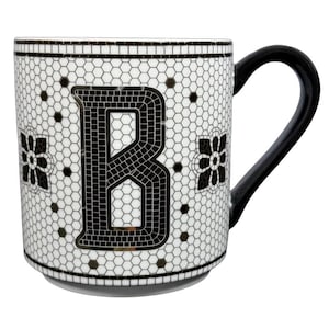 Pode incluir: Caneca de cerâmica branca com uma alça preta, apresentando um design em estilo mosaico. A caneca tem uma grande letra "B" preta no centro, rodeada por um padrão de favo de mel e detalhes florais. A borda e a base têm uma borda preta.
