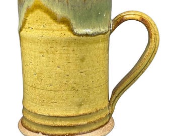 May include: A handmade ceramic mug with a large handle. The mug features a textured, cylindrical body with a light yellow glaze and a green rim. The base is a lighter, unglazed color.