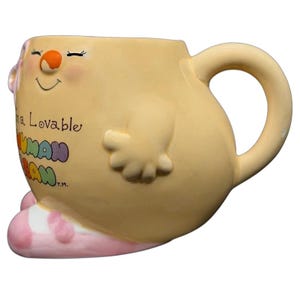Puede incluir: Una taza de cerámica amarilla caprichosa con una cara sonriente, un cuerpo redondeado y un asa curva. La taza presenta un diseño de dibujos animados con brazos, piernas y el texto "a Lovable HUMAN BEAN". Los pies son de rayas rosas y blancas.