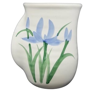Puede incluir: Jarrón de cerámica blanca con una forma curva única. Está adornado con flores de iris azules y hojas verdes pintadas a mano. El diseño es simple y elegante, adecuado para exhibir flores o como pieza decorativa.