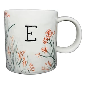 Könnte beinhalten: Weiße Keramiktasse mit dem schwarzen Buchstaben "E" und floralen Illustrationen in Orange, Grün und Blau. Die Tasse hat einen gebogenen Henkel und eine glatte Oberfläche. Das florale Design umschließt die Tasse.