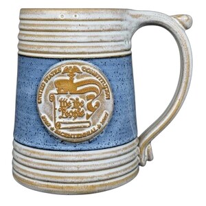 Può includere: Un boccale di birra in ceramica con finitura maculata blu e beige. Il boccale presenta un'ampia maniglia e un emblema circolare con le parole "United States Constitution" e "We the People".