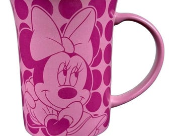 May include: Pink ceramic mug featuring Minnie Mouse. The mug has a pink polka dot pattern and a large image of Minnie Mouse. The handle is pink. The mug is a standard size for coffee or tea.
