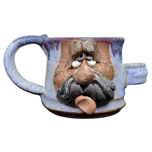 May include: Handmade ceramic mug with a sculpted face design. The mug features a blue and brown color scheme, with a handle and a small side tab. The face has a mustache, nose, and eyes, giving it a unique, artistic appearance.