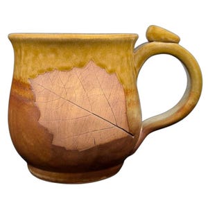Puede incluir: Taza de cerámica hecha a mano con una impresión de hoja. La taza presenta un esquema de color marrón y beige, con un gran diseño de hoja en la parte delantera. El asa tiene una forma redondeada única con una pequeña pieza de acento.