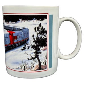 Op de afbeelding: Witte keramische mok met een winterse treinscène. De trein is zilver en rood, tegen een besneeuwd landschap met donkere bomen. De mok heeft een rode rand en een blauwe verticale streep. De mok heeft een oor.