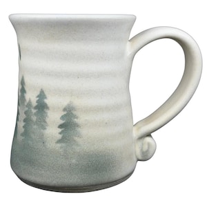 May include: A cream-colored ceramic mug with a forest scene painted in shades of green. The mug has a large handle and a slightly flared rim. The design features several stylized evergreen trees.