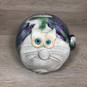 May include: A ceramic cat-shaped piggy bank. The bank features a white face with blue eyes and a round nose. The body is a mix of green, purple, and blue hues. The bank has a slot on top for inserting coins and a small tail.