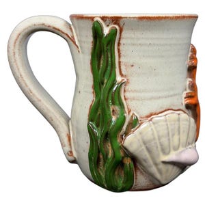 Puede incluir: Taza de cerámica color crema con borde y asa marrones. La taza presenta un diseño en relieve de algas verdes, una concha blanca y una estrella de mar naranja. Una pieza única de cerámica hecha a mano.