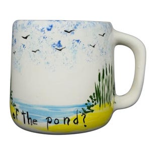 Puede incluir: Taza de cerámica blanca con un paisaje pintado a mano. El diseño incluye un cielo azul con pájaros, una costa amarilla y azul y juncos verdes. El texto "of the pond?" está escrito en la taza.