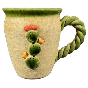 Op de afbeelding: Een beige keramische mok met een groene rand en een gedraaide handgreep. De mok heeft een 3D cactusontwerp met groene cactussen, oranje bloemen en gele accenten. De mok is ongeveer 10 cm hoog.