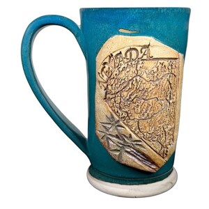 Può includere: Una tazza in ceramica turchese con un manico grande. La tazza presenta una mappa in rilievo color beige del Canada con la scritta "CANADA" in alto. La base della tazza è bianca.