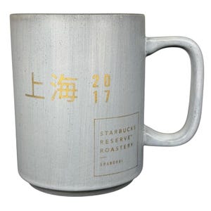 May include: A light gray ceramic mug with a large handle. The mug features gold text that reads "上海 2017" and a rectangular label with "STARBUCKS RESERVE ROASTERY SHANGHAI". The mug has a textured surface.