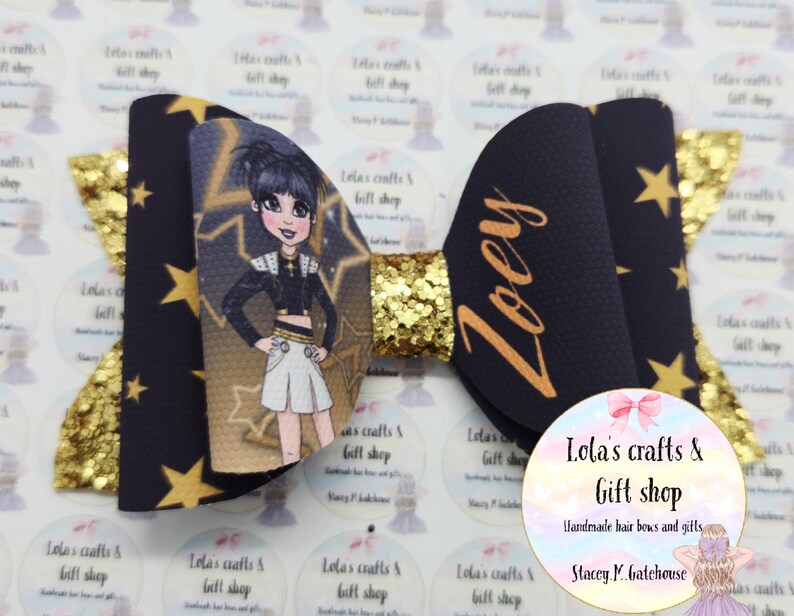 May include: A handmade hair bow featuring a cartoon character, gold glitter, and star accents. The bow is navy blue with gold stars and the name "Zoey" in gold script. The bow is from Lola's Crafts & Gift Shop.