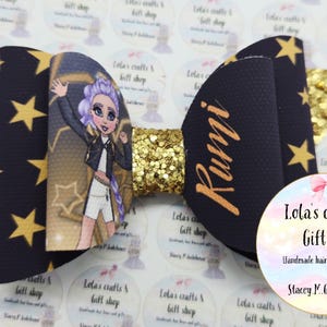 May include: A handmade hair bow with a black background and gold stars. The bow features a cartoon illustration of a girl with purple hair and the name "Rumi" in gold. A gold glitter center adds sparkle.