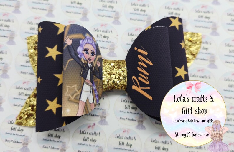 May include: A handmade hair bow with a black background and gold stars. The bow features a cartoon illustration of a girl with purple hair and the name "Rumi" in gold. A gold glitter center adds sparkle.