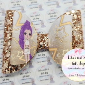 May include: A handmade hair bow featuring a cartoon girl with purple hair and gold stars on a white background. The bow has gold glitter accents and the text "Lola's crafts & gift shop" is visible.