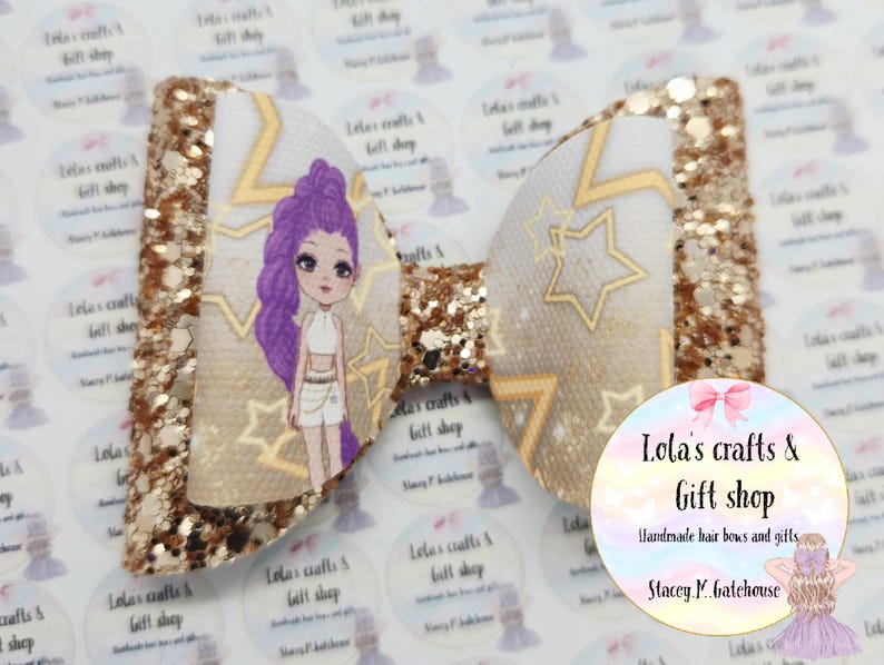 May include: A handmade hair bow featuring a cartoon girl with purple hair and gold stars on a white background. The bow has gold glitter accents and the text "Lola's crafts & gift shop" is visible.