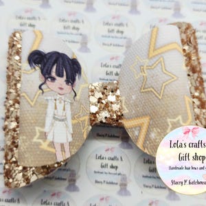 May include: A handmade hair bow with a glittery gold finish. The bow features a cartoon illustration of a girl in a white dress and gold stars. The bow is from Lola's Crafts & Gift Shop.