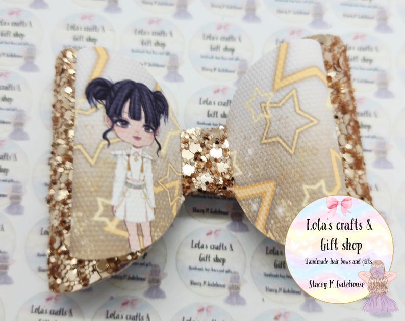 May include: A handmade hair bow with a glittery gold finish. The bow features a cartoon illustration of a girl in a white dress and gold stars. The bow is from Lola's Crafts & Gift Shop.