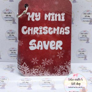 May include: A red, rectangular Christmas-themed item with the text "My Mini Christmas Saver" in white. It has a silver ring and snowflake decorations. The background features repeating circular stickers with text.