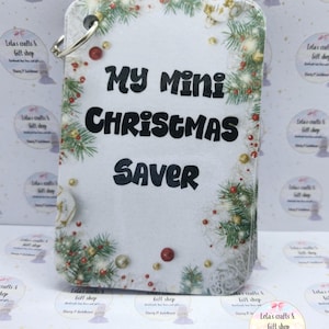 May include: A rectangular Christmas gift tag with the words "My Mini Christmas Saver" in bold black. It features a festive border of green fir, red berries, and gold baubles. A silver ring is attached at the top for hanging. The tag is white.
