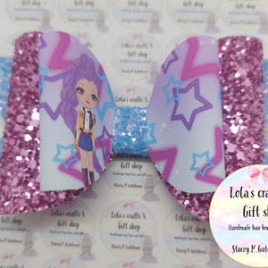 May include: A handmade hair bow featuring a cartoon character with purple hair and a star pattern. The bow has a glittery pink center and light blue accents. The text "Lola's crafts & Gift shop" is visible.