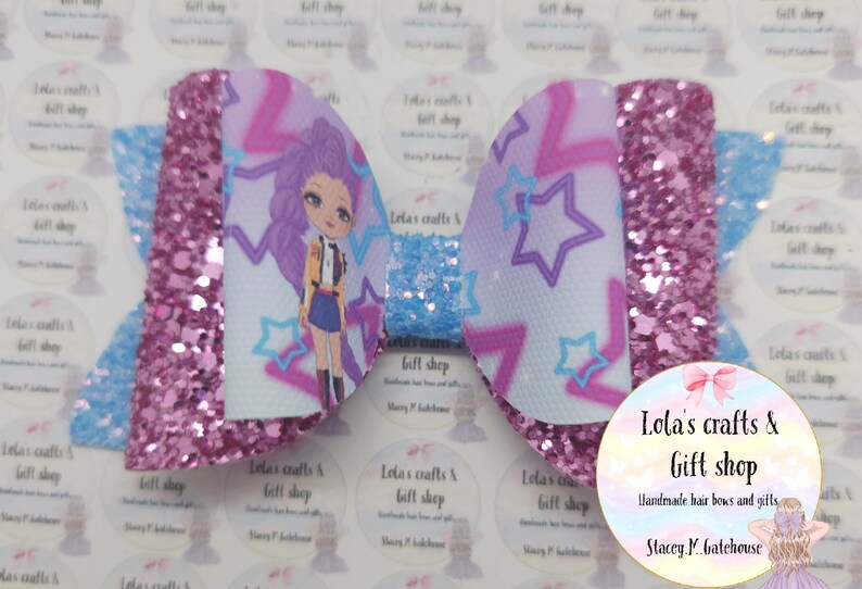 May include: A handmade hair bow featuring a cartoon character with purple hair and a star pattern. The bow has a glittery pink center and light blue accents. The text "Lola's crafts & Gift shop" is visible.