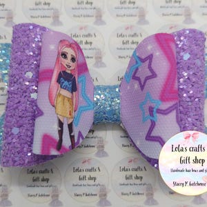 May include: A large, decorative hair bow featuring a cartoon girl with pink hair and a blue top. The bow has a purple glittered texture on the sides and a light blue center. The design includes stars and the text "Lola's crafts & Gift shop".