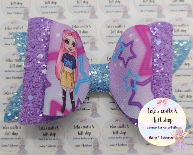 May include: A large, decorative hair bow featuring a cartoon girl with pink hair and a blue top. The bow has a purple glittered texture on the sides and a light blue center. The design includes stars and the text "Lola's crafts & Gift shop".