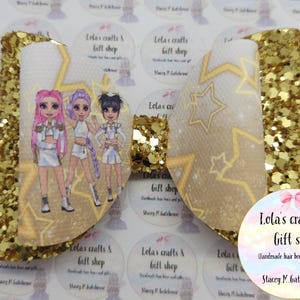 May include: A large, gold glitter hair bow with a white center featuring a cartoon image of three girls. The bow has a star pattern and is labeled "Lola's crafts & Gift shop".