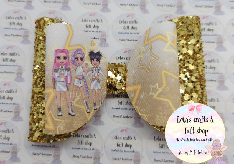 May include: A large, gold glitter hair bow with a white center featuring a cartoon image of three girls. The bow has a star pattern and is labeled "Lola's crafts & Gift shop".