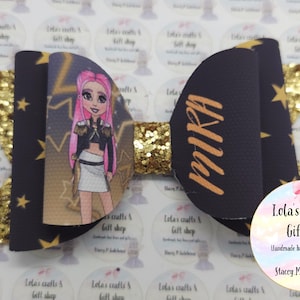 May include: A handmade hair bow featuring a cartoon character with pink hair. The bow has a black background with gold stars and the name "MIRA" in gold. The center is gold glitter.