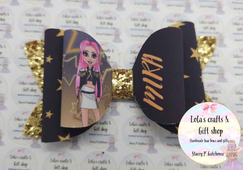 May include: A handmade hair bow featuring a cartoon character with pink hair. The bow has a black background with gold stars and the name "MIRA" in gold. The center is gold glitter.