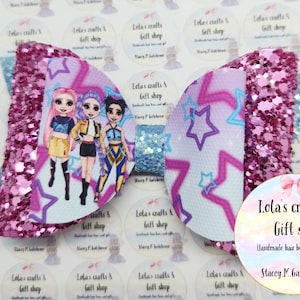 May include: A large, handmade hair bow with a glittery pink and blue design. The bow features a central panel with a cartoon illustration of three girls and a star pattern. The bow is from Lola's Crafts & Gift Shop.