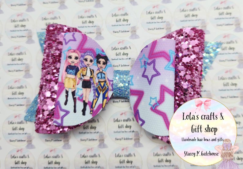 May include: A large, handmade hair bow with a glittery pink and blue design. The bow features a central panel with a cartoon illustration of three girls and a star pattern. The bow is from Lola's Crafts & Gift Shop.