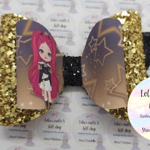 May include: A handmade hair bow with a cartoon girl design. The bow features a gold glitter accent, a black center, and a printed design with stars and a cartoon girl with pink hair. The bow is from Lola's Crafts & Gift Shop.