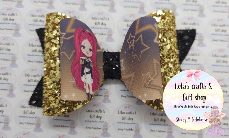 May include: A handmade hair bow with a cartoon girl design. The bow features a gold glitter accent, a black center, and a printed design with stars and a cartoon girl with pink hair. The bow is from Lola's Crafts & Gift Shop.