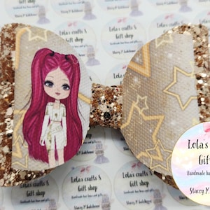 May include: A large, handmade hair bow with a glittery gold and tan design. One side features a cartoon girl with pink hair, while the other side has a star pattern. The bow is from Lola's Crafts & Gift Shop.