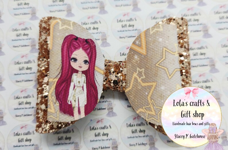 May include: A large, handmade hair bow with a glittery gold and tan design. One side features a cartoon girl with pink hair, while the other side has a star pattern. The bow is from Lola's Crafts & Gift Shop.