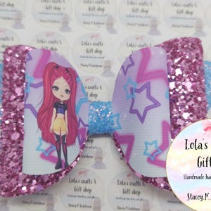 May include: A large, decorative hair bow featuring a cartoon girl design. The bow has a glittery pink center and edges, with a light blue center and wings. The bow has a white background with purple stars and a cartoon girl. The bow is from Lola's Crafts & Gift Shop.