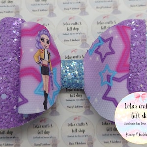 May include: A large, handmade hair bow with a glittery purple and light blue design. The bow features a cartoon girl graphic and star patterns. The bow is from Lola's Crafts & Gift Shop.