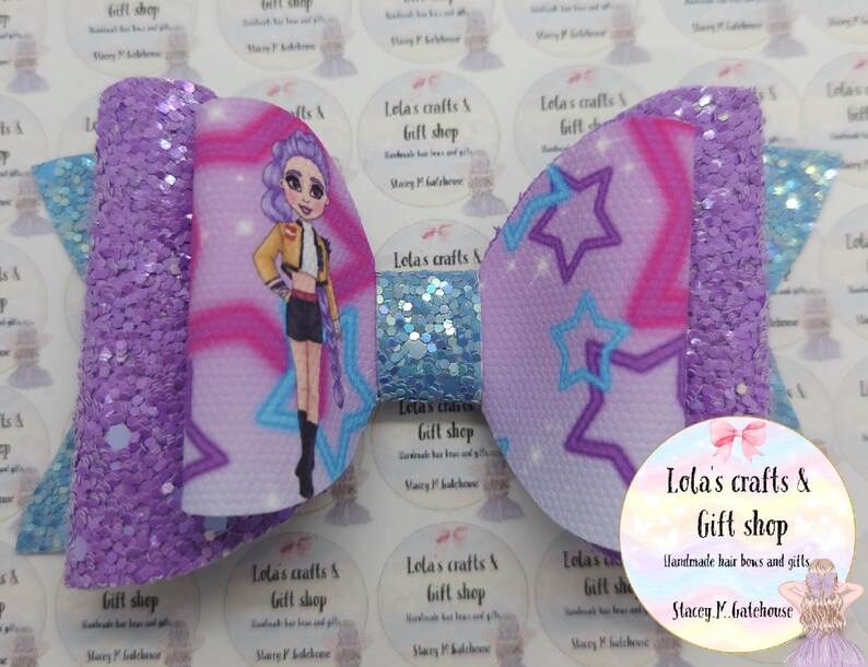 May include: A large, handmade hair bow with a glittery purple and light blue design. The bow features a cartoon girl graphic and star patterns. The bow is from Lola's Crafts & Gift Shop.